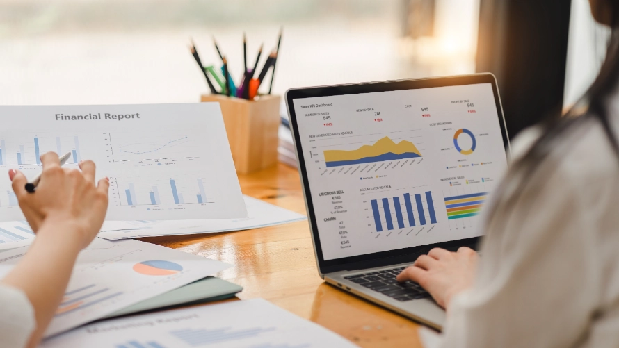 Close-up: Hands typing on a laptop showing a sales KPI dashboard. Financial printouts and a pen holder surround it on a wooden desk.